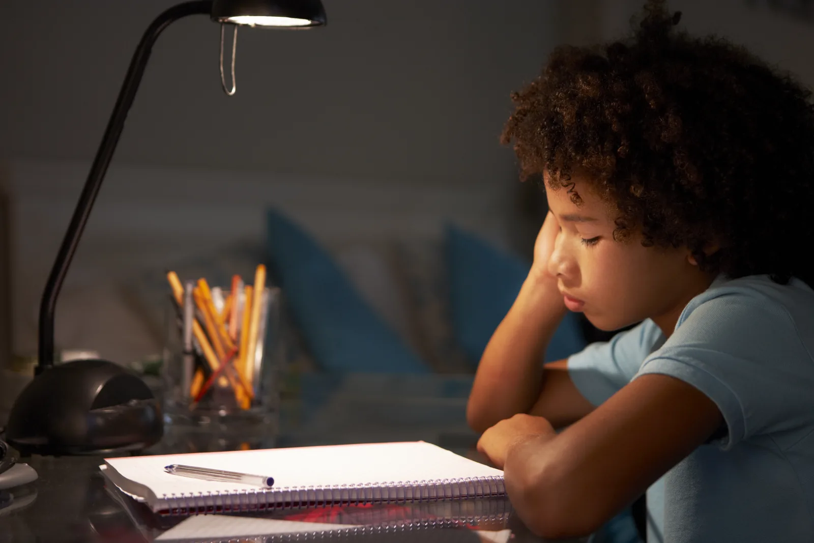 Boy looking tired while doing homework under a desk lamp
